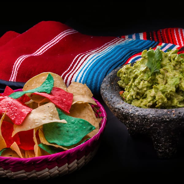 Colorful tortilla chips in a basket next to a molcajete filled with guacamole, on a striped blanket.