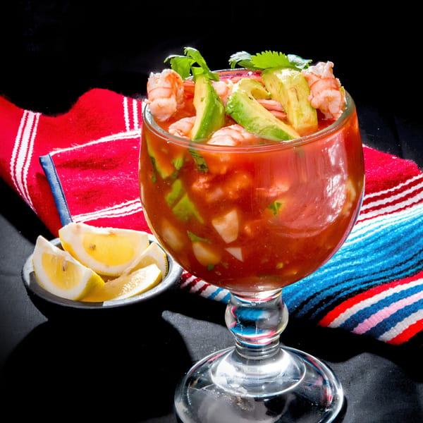 Las Tres Casuelas' shrimp cocktail with avocado, cilantro, and lemon wedges on a colorful striped cloth.