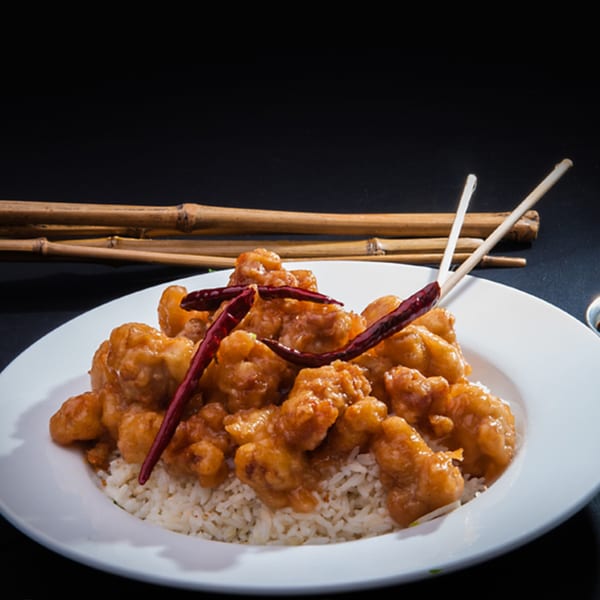 Orange chicken with red chili peppers and white rice on a white plate with chopsticks.