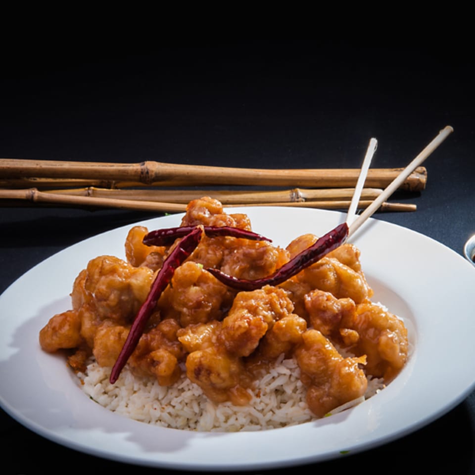 Orange chicken with red chili peppers and white rice on a white plate with chopsticks.