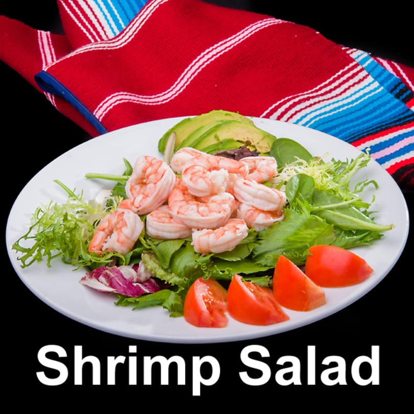 A white plate of shrimp salad with mixed greens, avocado slices, and tomato wedges, on a black background.